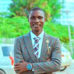 Professional portrait of a man wearing a brown checkered suit, white shirt, and patterned tie stands outdoors with his arms crossed, smiling at the camera, with greenery and parked cars in the background.