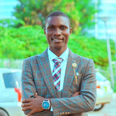 Professional portrait of a man wearing a brown checkered suit, white shirt, and patterned tie stands outdoors with his arms crossed, smiling at the camera, with greenery and parked cars in the background.